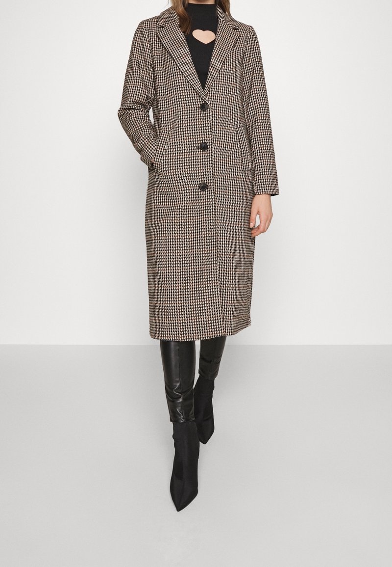 Manteau long à motif pied-de-poule en noir, beige et marron avec revers crantés et trois boutons noirs. Associé à un pantalon en cuir noir et des bottes à talons.