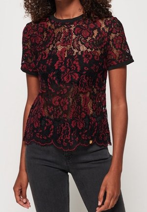 Kobieta ubrana w dopasowaną czarno-czerwoną koronkową bluzkę w kwiaty z krótkim rękawem, w parze z ciemnoszarymi dżinsami, stojąca z rozluźnionymi ramionami.