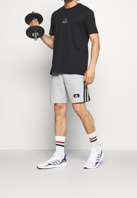 Camiseta de algodón negra, pantalones cortos grises con acentos negros, zapatillas blancas con detalles en azul y rojo, sosteniendo una mancuerna negra.