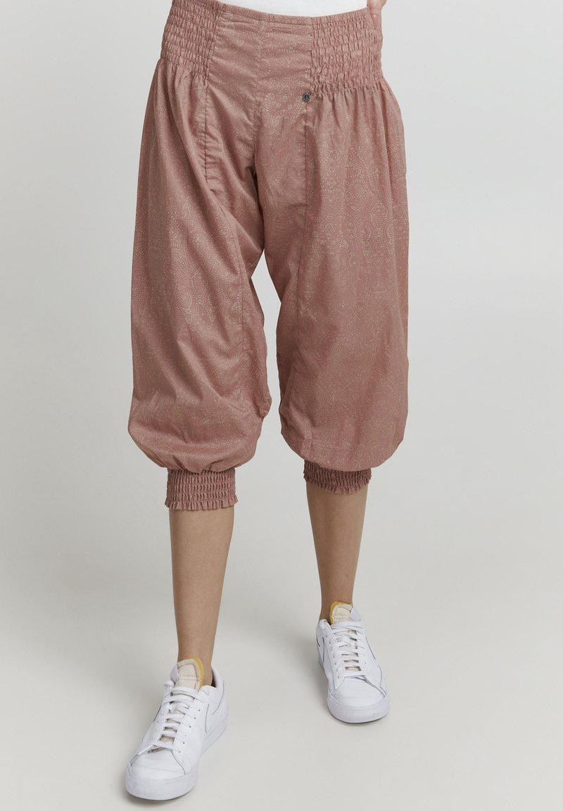 Pantalon court ample rose clair avec taille et poignets élastiques, porté avec des baskets blanches à lacets sur un fond neutre.