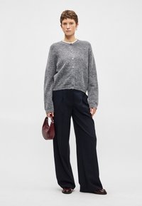 Grijze wollen cardigan met ronde hals en knoopsluiting, gedragen over marineblauwe pantalon met streepjes en wijde pijpen. Model houdt een kleine bordeauxrode handtas vast.