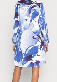 Osoba v modrých, fialových a bílých abstraktních květinových šatech s dlouhým rukávem, zobrazená zezadu na jednobarevném světlém pozadí.