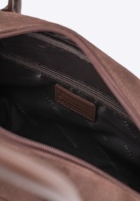 Interior de un bolso de ante marrón, con un bolsillo con cremallera y un forro de tela oscura con un sutil patrón de logo. Etiqueta de cuero marrón en el interior.