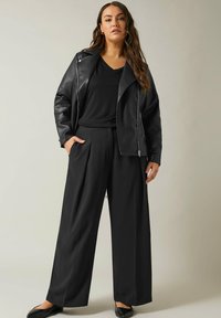 Zwart leren motorjack met zilveren ritssluiting, zwarte t-shirt en ruime zwarte broek. Model draagt zwarte flats en heeft de handen in de zakken.