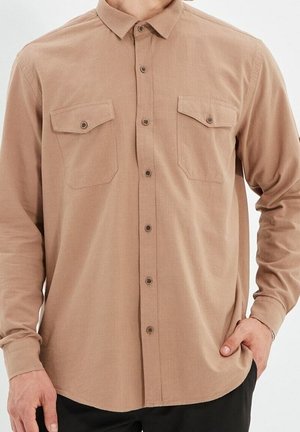 Mand iført en beige langærmet skjorte med to brystlommer med knapper, hånd hviler i lommen på sorte bukser.