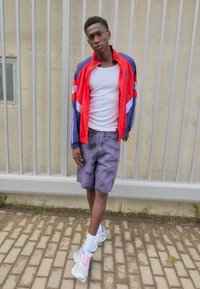 Crvena i plava jakna na zip preko bijele majice bez rukava, uparena s ljubičastim šortsem u tie-dye stilu i sivim tenisicama s ružičastim naglascima. Stoji na asfaltu.