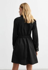 Abito nero con righe verticali, maniche lunghe, vita con cintura e polsini con bottoni, caratterizzato da un taglio ampio fino al ginocchio e una texture morbida.