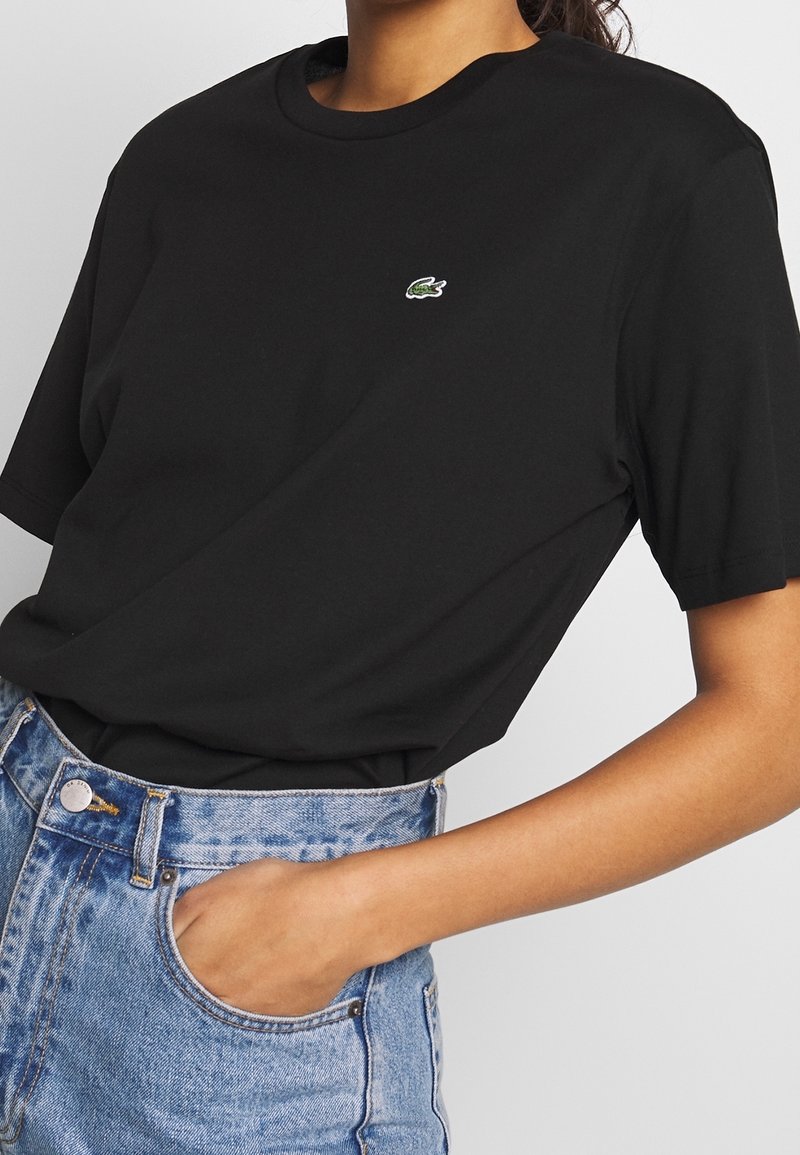 Schwarzes T-Shirt mit kurzen Ärmeln aus Baumwolle und Rundhalsausschnitt, mit einem kleinen gestickten grünen Krokodil-Logo auf der Brust.