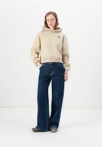 Felpa beige corta con tasca frontale e logo, abbinata a jeans blu larghi e sneakers grigie, su uno sfondo semplice.
