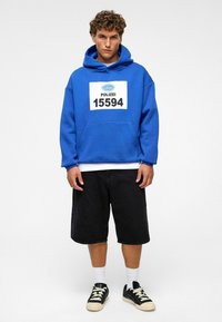 Blauwe hoodie met een logo en "POLIZEI 15594" gedrukt op de voorkant; gecombineerd met zwarte shorts en zwarte sneakers met witte zolen.