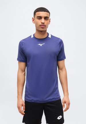 Jonge man staand, gekleed in een blauw sportshirt en zwarte korte broek met sportmerkenlogo's op een effen witte achtergrond.