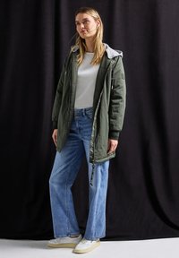 Oliivinvihreä hupparitakki, jossa on harmaa vuori, pitkät hihat ja vetoketjulla varustettu etuosa, yhdistettynä vaaleansinisiin leveälahkeisiin farkkuihin ja valkoisiin kenkiin.