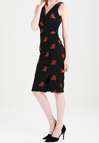 Kobieta ubrana w czarną, bezrękawnikową sukienkę do kolan w czerwone róże, z rozcięciem z przodu, zestawioną z czarnymi szpilkami.