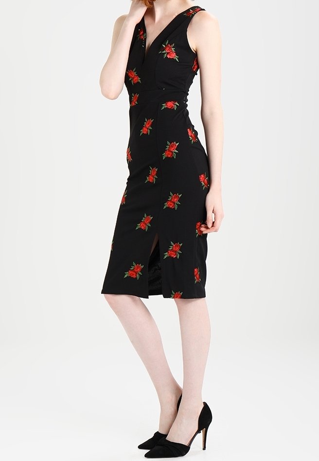 Kobieta ubrana w czarną, bezrękawnikową sukienkę do kolan w czerwone róże, z rozcięciem z przodu, zestawioną z czarnymi szpilkami.