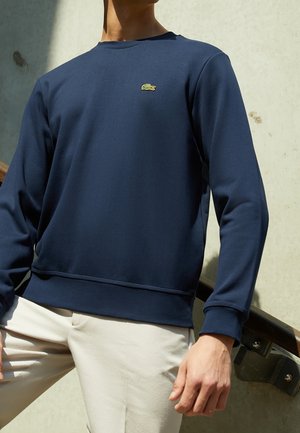 Mężczyzna ubrany w granatową koszulę z długim rękawem z małym zielonym logo krokodyla i jasnobeżowe spodnie, stojący obok poręczy schodów.