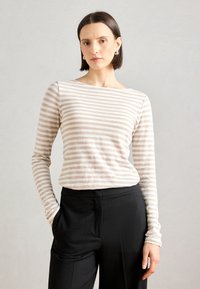 Langermet topp i lys beige med horisontale striper, med båt-halsdesign. Parret med svarte bukser, står modellen med en hånd i lommen.