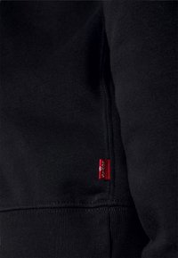 Gros plan sur un tissu noir avec une petite étiquette rouge Levi’s cousue près de la couture sur la couture latérale d’un vêtement.