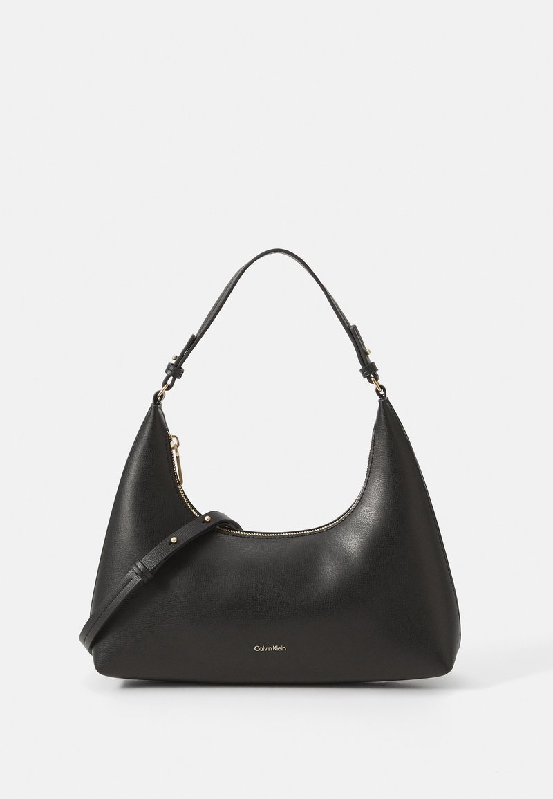 Sac à main en cuir noir Calvin Klein avec poignée supérieure, bandoulière réglable et fermeture éclair dorée sur fond clair.