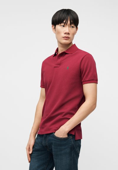 Νέος άνδρας που στέκεται με το χέρι στην τσέπη του τζιν, φορώντας κόκκινο πουκάμισο πόλο, μπροστά σε απλό φωτεινό φόντο, κοιτάζοντας προς τα πλάγια.