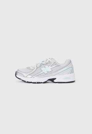 Witte en zilveren sport sneaker met een lichtblauwe "N"-logo, mesh lagen en witte veters op een lichtgrijze achtergrond.