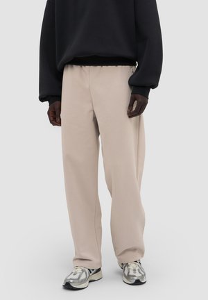 Man draagt beige wijde broek, zwarte longsleeve trui en wit-grijze sneakers, staand tegen een effen lichtgrijze achtergrond.