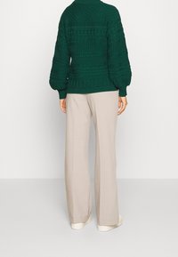Pull vert tricoté épais avec des motifs texturés, associé à un pantalon large beige clair et des chaussures blanches.