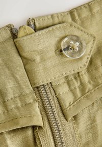 Gros plan sur un pantalon en tissu beige montrant un bouton clair et une fermeture éclair partiellement fermée avec un tirette beige marquée C5.