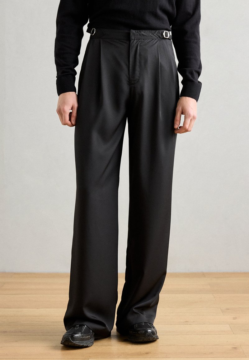 Pantalones negros de pierna ancha con una textura suave, que presentan doble hebilla lateral y un diseño de pliegues en la parte delantera. Combinados con zapatos negros.