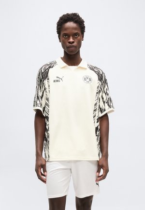 Young man standing wearing white Borussia Dortmund football shirt with black abstract side patterns and matching white shorts.