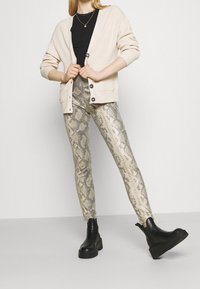 Beige ribbad cardigan med svarta knappar, bärs över en svart topp, i kombination med metallisk ormskinnstrykt leggings och kraftiga svarta ankelskor.
