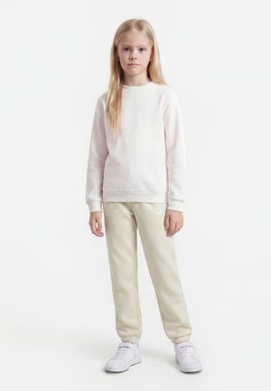 Junges Mädchen mit langen blonden Haaren steht gerade, trägt einen hellen Pullover, beige Jogginghosen und weiße Turnschuhe vor weißem Hintergrund.
