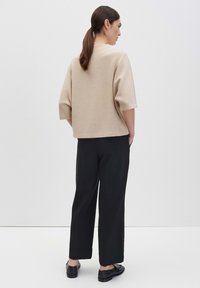 Frau mit zurückgebundenem dunklem Haar, die ein beigefarbenes Kurzarmoberteil, schwarze weite Hosen und schwarze Schuhe trägt, steht vor einem weißen Hintergrund.