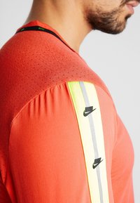 Czerwona koszulka sportowa z teksturowanym wykończeniem, z czarnym logo Nike oraz neonowo-żó�łtymi i szarymi paskami na ramieniu.