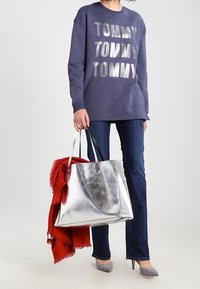 Borsa tote metallizzata argento con due manici, abbinata a una felpa blu navy con grandi scritte argentate, jeans blu scuro e tacchi grigi. Una sciarpa rossa è appoggiata sulla borsa.