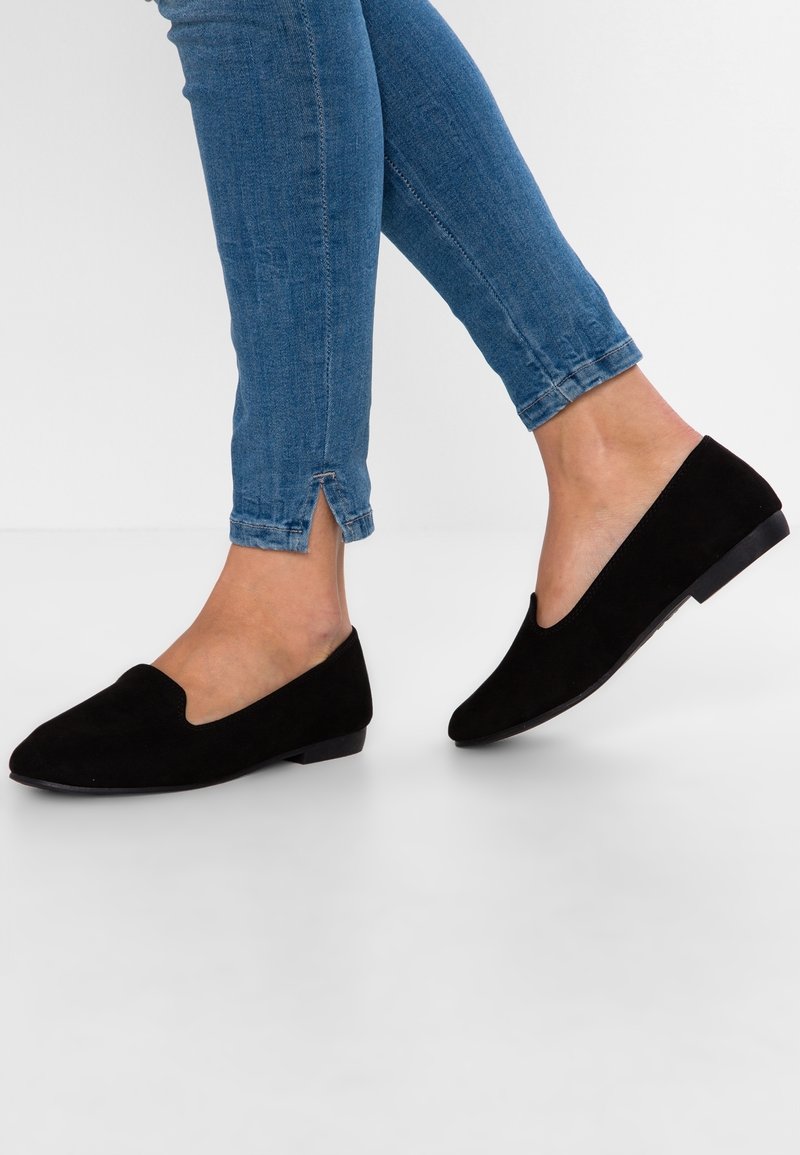 Pieds portant des chaussures plates noires à enfiler associées à un jean skinny bleu, debout sur une surface blanche.
