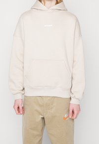 Personne portant un sweat à capuche beige oversize avec le texte "WRSTBHVR" et un pantalon Nike beige avec un logo swoosh orange, debout devant un fond uni.