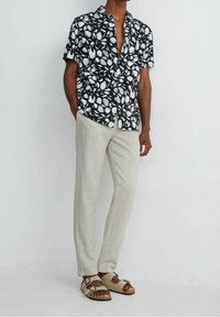 Chemise noire à manches courtes avec un motif de feuilles blanches, associée à un pantalon en lin beige clair et des sandales brunes à deux brides et boucle.