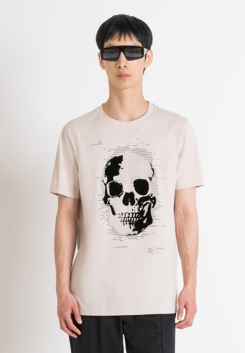Camiseta de algodón beige con un gráfico de calavera negra y distorsiones abstractas. Modelo que lleva gafas de sol rectangulares negras y pantalones oscuros.