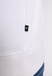 Sweatshirt blanc en coton avec ourlet côtelé et coutures latérales, comportant une petite étiquette logo noire sur le côté gauche près du bas.