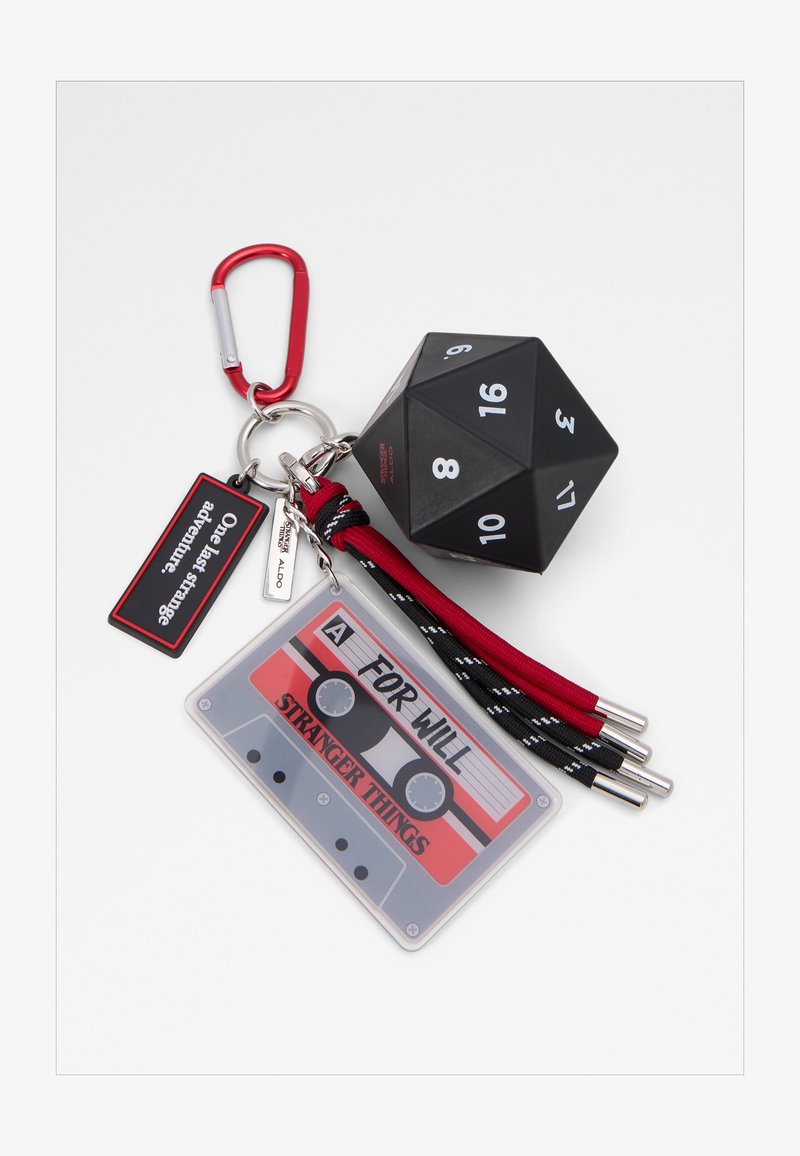 Fermoir de porte-clés rouge tenant un dé noir à 20 faces, une étiquette de cassette audio étiquetée "POUR WILL Stranger Things" et une étiquette "Une dernière aventure étrange".