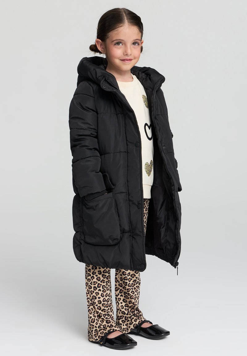 Czarna kurtka puchowa z kapturem, z dużymi przednimi kieszeniami, zestawiona z kremowym swetrem i spodniami w panterkę. Czarne lakierowane buty dopełniają look.