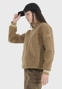 Braune Fleecejacke mit hohem Kragen, langen Ärmeln und weißen gestreiften Bündchen. Kombiniert mit olivgrünen Hosen und einer schwarzen Mütze.