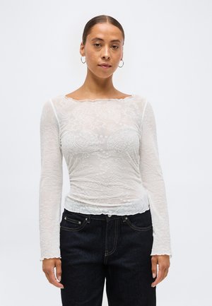 Ženska z zategnjenimi lasmi, ki nosi obročaste uhane, belo prosojno majico z vzorcem paisley z dolgimi rokavi in temne kavbojke, na enobarvnem ozadju.