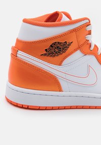 Valkoisesta ja kirkkaan oranssista synteettisestä nahasta valmistettu puolikorkea tennari, jossa on valkoiset kengännauhat, kuvioitu pohja ja musta Air Jordan -logo.