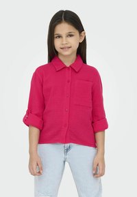 Camisa magenta de botones con cuello, mangas largas subidas, tejido texturizado y un bolsillo en la parte frontal, combinada con jeans de mezclilla azul claro.