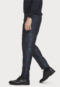 Mężczyzna idący, w granatowych spodniach w turkusowy wzór palmy, czarnych butach wizytowych i czarnych skarpetach na białym tle.