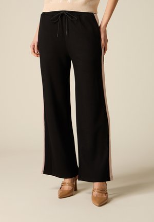 Vrouw draagt zwarte broek met wijde pijpen en beige zijstrepen, beige mouwloze top en beige spitse hoge hakken met bandjes.