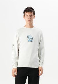 Lange mouwen, lichtgrijze katoenen t-shirt met een blauwe hondenafbeelding op de borst. Tekst afgedrukt op de linker mouw. Zwarte broek gedragen.
