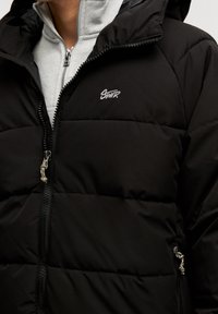 Chaqueta negra tipo puffer con diseño acolchado, que incluye una cremallera frontal, capucha ajustable y un logo bordado en el pecho. Textura suave.