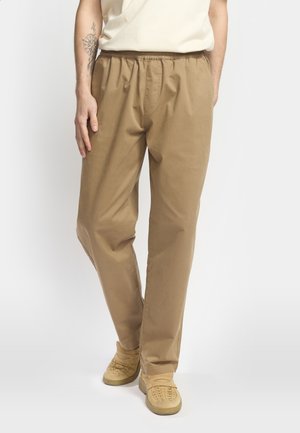Pantaloni beige con vita elasticizzata e design a gamba dritta, realizzati in tessuto leggero. Indossati con sneakers color tan, caratterizzate da una texture a coste.